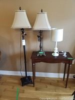 Three lamps: one tall black floor lamp with square base and fabric shade, one matching table lamp on wooden table with fabric shade, small white plastic lamp with cylindrical fabric shade on same table.