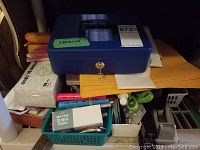 Blue metal lockable cash box with key on top of assortment of office supplies including envelopes, stapler, and scissors in baskets and boxes.