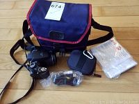Fujifilm Finepix digital camera with black strap, manual, CD, and blue and red camera carrying case on hardwood floor.