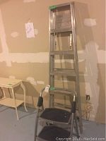 Photo shows a six-foot aluminum step ladder against a wall with two small tables nearby and an electrical cord on the floor.