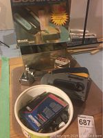 Lot with Bostik 260 electric stapler in box, Everbuilt manual staple gun, metal tins, pack of staples, bucket with staples, and candle tin on table.