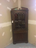 Front view of wood corner cabinet with glass door and decorative lattice, showing interior shelving, drawer and lower doors.