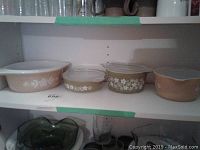 Four vintage Pyrex casserole dishes on shelf, three with lids, showing peach and olive green colors with floral patterns.