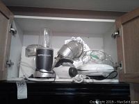NutriBullet blender with blending cup, white food processor, Sunbeam Mix Master stand mixer, Salton electric grill, and a silver wire basket in a cupboard shelf