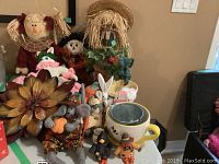 A collection of seasonal decorations including scarecrow dolls, bunny figurines, a large yellow ceramic mug, and metal flower wall art all arranged on a table surface.