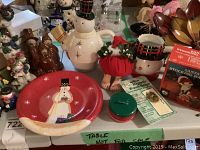 Photo showing snowman teapot missing button nose, red snowman plate, snowman mug, red fabric Christmas figure, and boxed Christmas decorations on table