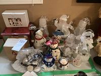 Full collection of assorted snowmen figurines in ceramic and clear acrylic, some designed as tea light holders, with two storage boxes and a bag of white tea lights on display on a tabletop.