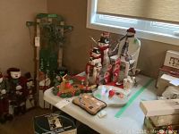 Wooden snowmen statues, rocking chair with snowman figure, Santa faces, reindeer head, and various wooden holiday decorations arranged on a table in front of a window.