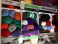 Photo showing skeins of yarn in three white plastic baskets placed on a shelf, displaying a variety of colors including reds, greens, blues, and purples.