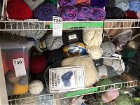 Photo showing various skeins of yarn in multiple colors on metal shelving with a label indicating yarn and brand tags visible.