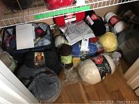 Yarn skeins in multiple colors and brands stored on a closet floor, including Red Heart and large black skein with label showing person in orange scarf. Partially used skeins and bags with yarn visible.