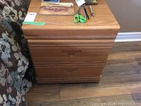 Set of two wooden night stands with two drawers each, seen side by side on a wooden floor. The stands have a medium brown finish and simple V-shaped drawer handles.