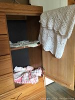 Knitted baby sweaters and hats stored inside a wooden cabinet with solid white crocheted baby blanket hanging on the door