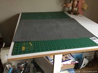 Photo of two hobby cutting mats stacked on a table. The bottom mat is green with yellow grid and measures around 24x18 inches. The top mat is gray with orange grid, larger at approximately 23.5x35.5 inches, showing signs of wear.