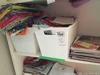 View of two white plastic bins filled with assorted fabric pieces, some folded and showing various colorful prints including owls and geometric designs. Also visible is a shelf with stacked magazines.