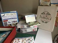 Photo showing various cross stitch supplies including large book, clear plastic bins with threads and embroidery materials, finished embroidery on fabric stretched on a board.