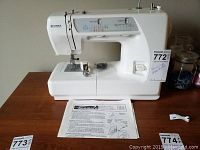 Front and side view of the white Kenmore sewing machine showing stitch selector panel, control knobs, and instruction sheet underneath the machine.