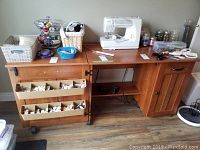 Front view of wooden sewing cabinet with sewing machine on top. Cabinet has multiple baskets and containers for sewing supplies but these are not included in the lot.