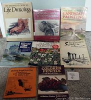 Seven art instruction books arranged on floor, showing covers and titles related to life drawing, landscape painting, watercolor, flower painting, and drawing techniques.