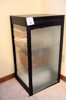 Black display storage cabinet with frosted glass front door, closed, showing contents inside through clear side panels including boxes and baskets.