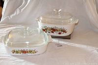 Two white glass casserole dishes with L'Echalote vegetable pattern, each with clear glass lid, on white backdrop