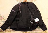 Front view of black ladies motorcycle jacket, partially unzipped showing quilted liner inside, adjustable waist strap visible