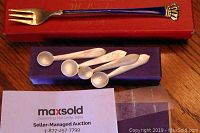 Set of four small sterling silver salt spoons and one Denmark sterling pickle fork with blue enamel handle displayed on table with boxes and MaxSold card.