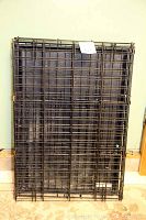 Collapsed metal wire grid pet cage with a black plastic tray underneath, placed on carpeted floor against a wall.
