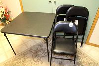 Black rectangular card table with folding legs positioned next to three stacked black folding chairs with padded seats and backs, set on a beige patterned rug.