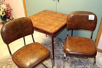 Photo showing brown card table with laminate top and two matching brown vinyl upholstered chairs with chrome legs on a patterned rug.