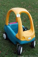 Front and side view of Little Tikes Cozy Coupe with yellow roof, blue body, and black steering wheel. Pink rumble seat at rear for doll placement. Plastic wheels and faded colors from sun.