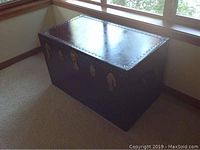 Front side view of the brown leather bound trunk with brass locks and trim, positioned next to a window on carpeted floor.
