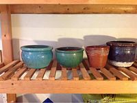 Four ceramic pots in turquoise blue, green, reddish-brown terracotta, and deep purple glaze on wooden shelf