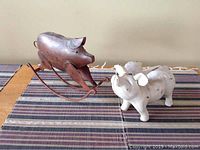 Two metal pig sculptures on a striped cloth surface, showing the rust finish rocking pig on metal rockers and the white flying pig bank.