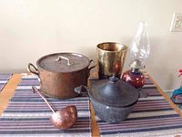 Overview photo of all items on table showing large brass copper pot with lid, dark grey pot with lid, brass vase, oil lamp, and ladle.