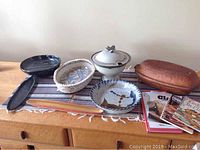 Photo showing six ceramic serving pieces on a wooden surface including a black oval dish, a woven style dish, a covered pot with lid, a round dish with design, a striped serving piece, and a large clay cooking pot with lid; also partially visible four cookbooks.