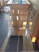A clear acrylic rolling bar cart with chrome handles and casters on slate floor.