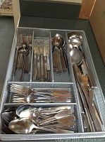 Metal mesh drawer organizer filled with various stainless steel flatware pieces including forks, knives, spoons, and serving spoons arranged in compartments.