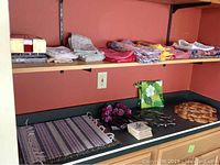 Counter with folded cloth napkins in various colors and patterns, decorative napkin rings shaped like flowers, round braided trivets, striped woven placemats with fringed edges, and small square coasters or napkins with floral patterns.