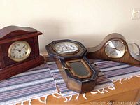 Wide shot showing all three clocks on a striped cloth-covered table: two mantle clocks and one wall clock with pendulum.
