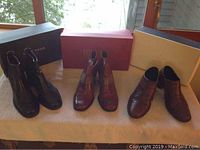 Three pairs of boots and leather ankle shoes on white cloth by window, shoeboxes behind