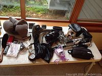 Wide view of all items on table including massager, hair dryers, curling irons, headphones, pillow, mirror, brushes, clocks, and carry cases