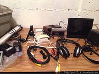 Photo showing assorted electronic items on wooden table including cables, black headphones, power adapters and black box device with dials