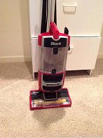 Front view of red Shark upright vacuum with Lift-Away canister and clear dust compartment on beige carpet.