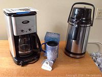 Photo showing Cuisinart coffee maker, Tiger thermos, and blue travel mug with snowflakes on wooden table.