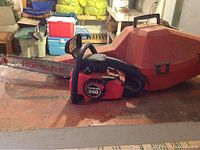 Red and black Homelite 240 chainsaw sitting on a surface with orange carrying case behind it, in indoor storage area.