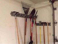 Long handled garden tools including rakes, shovels, and hoes hung on wall-mounted rack