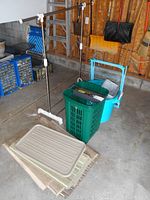 Wide shot of clothes rack, blue plastic pail, green laundry hamper with paper shredder on top, drying rack, rubber mats and some folded items on floor