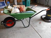 Green Fort brand wheelbarrow with orange-handled grips, black metal frame, and a green basin. Several garden tools and items are inside the wheelbarrow.