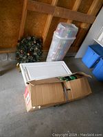 Storage bin containing Christmas bows and wrapping paraphernalia next to the wreath with rolls of wrapping paper in a plastic tall container.